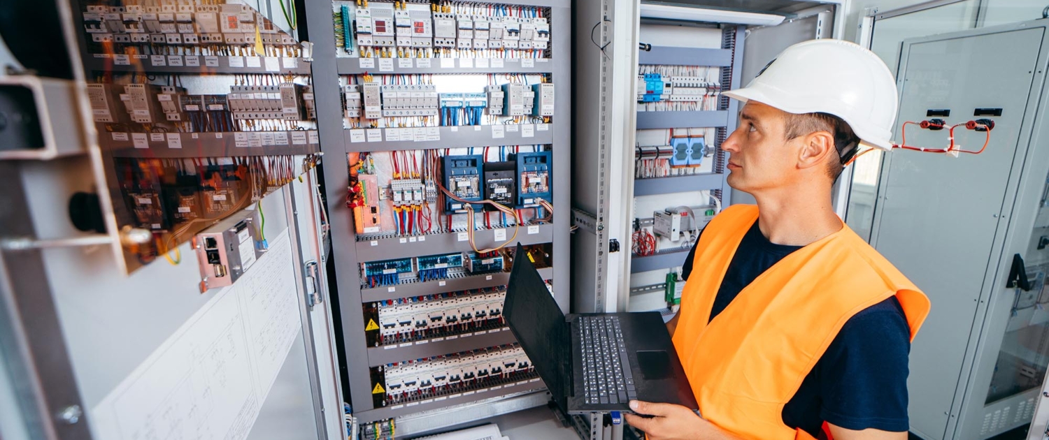 worker holding computer while looking over electrical switches