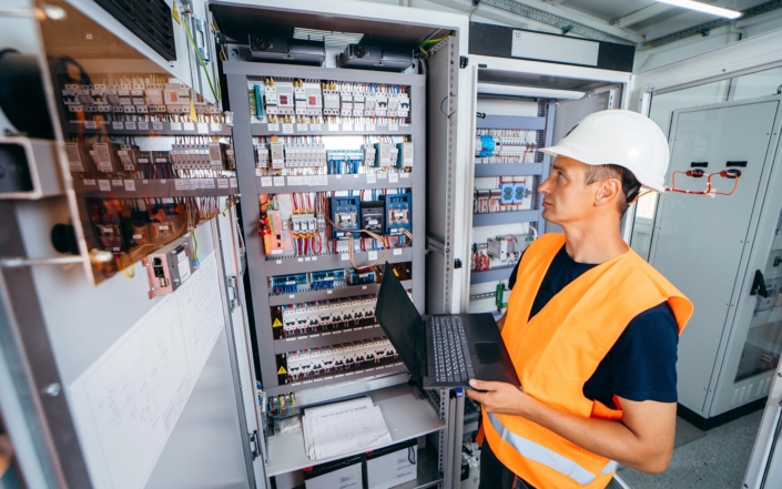 worker holding computer while looking over electrical switches